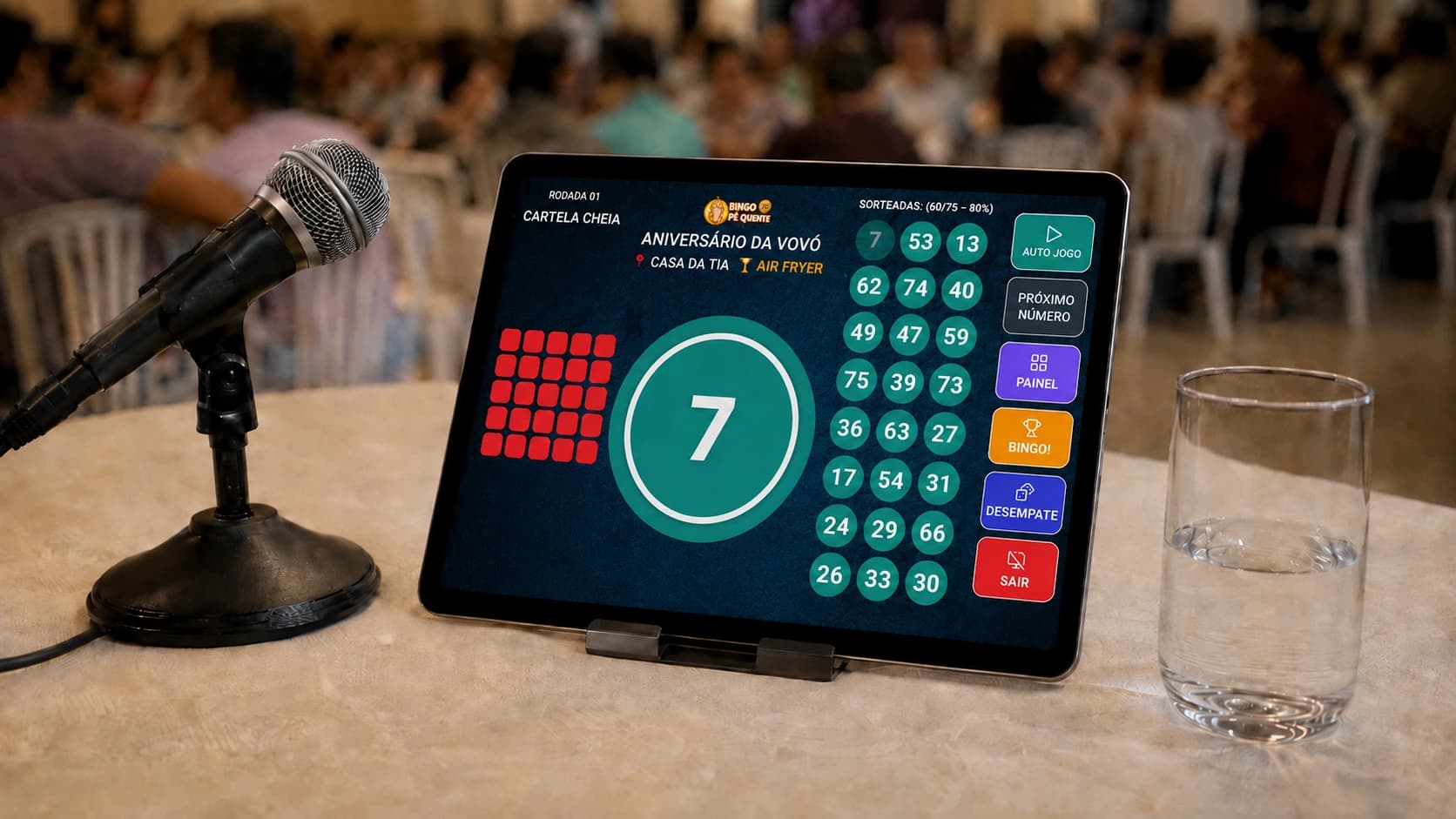 Mesa de controle do cantador em um bingo comunitário: microfone em pedestal, copo de água ao alcance da mão e um tablet apoiado mostrando a tela do sorteador do Bingo Pé Quente, com o número 7 sorteado em destaque, o padrão da rodada em vermelho, as bolas já cantadas e os botões de auto jogo e painel
