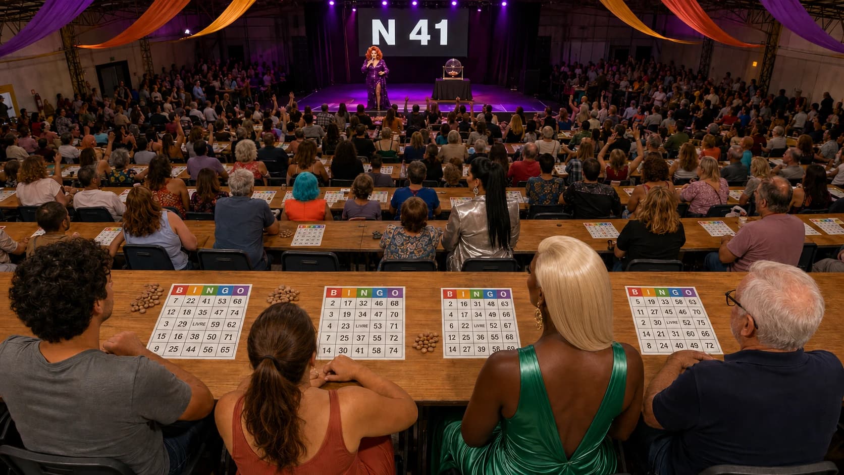Vista do fundo de um galpão comunitário lotado durante uma noite de bingo: centenas de pessoas de todas as idades sentadas em mesas compridas, cada uma com sua cartela de cabeçalho colorido B-I-N-G-O e montinhos de feijão, muitas com a mão erguida; entre o público, pessoas de vestido de festa, peruca platinada e brincos grandes acompanham sentadas; ao fundo, o palco iluminado com uma drag queen ao microfone, o globo de bingo e um telão exibindo N 41